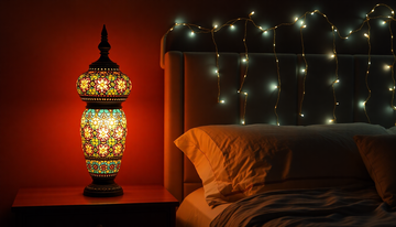 A glowing colorful Turkish mosaic glass table lamp on a nightstand beside a strand of fairy lights draped over a headboard