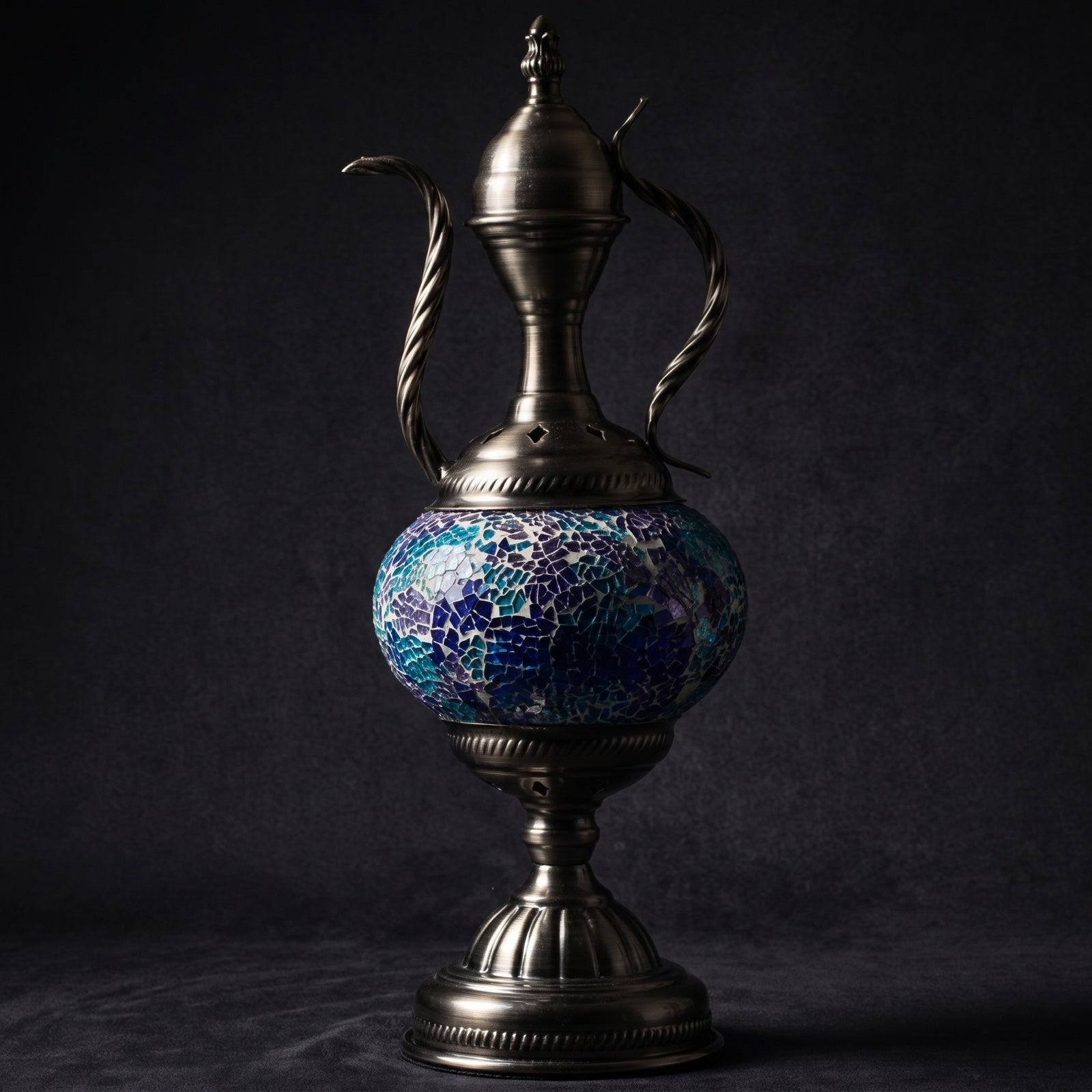 Unlit mosaic glass ewer-shaped table lamp showing intricate stained-glass tile pattern on a dark surface